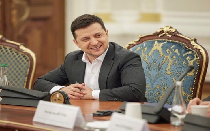 Volodymyr Zelenskyy smiling in a grey-black for mal coat over a plane white shirt.